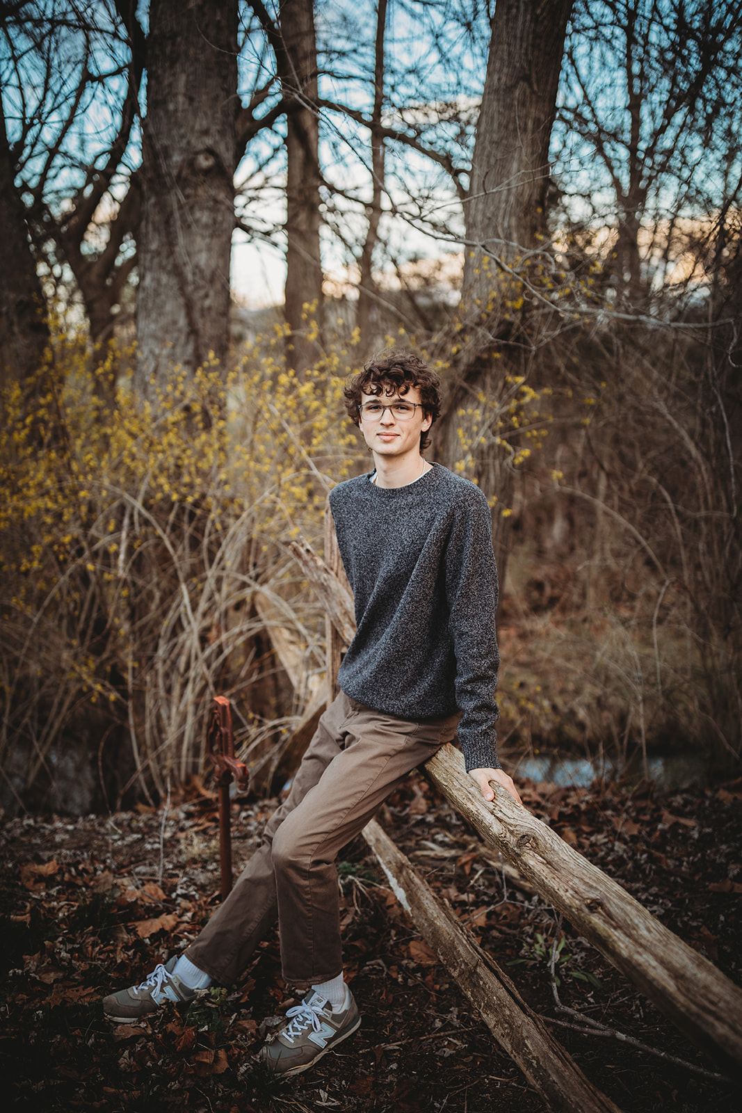 A teenage boy sits on a wooden fence by a creek in a grey sweater and tan pants after finding a Tuxedo Rental Harrisonburg, VA