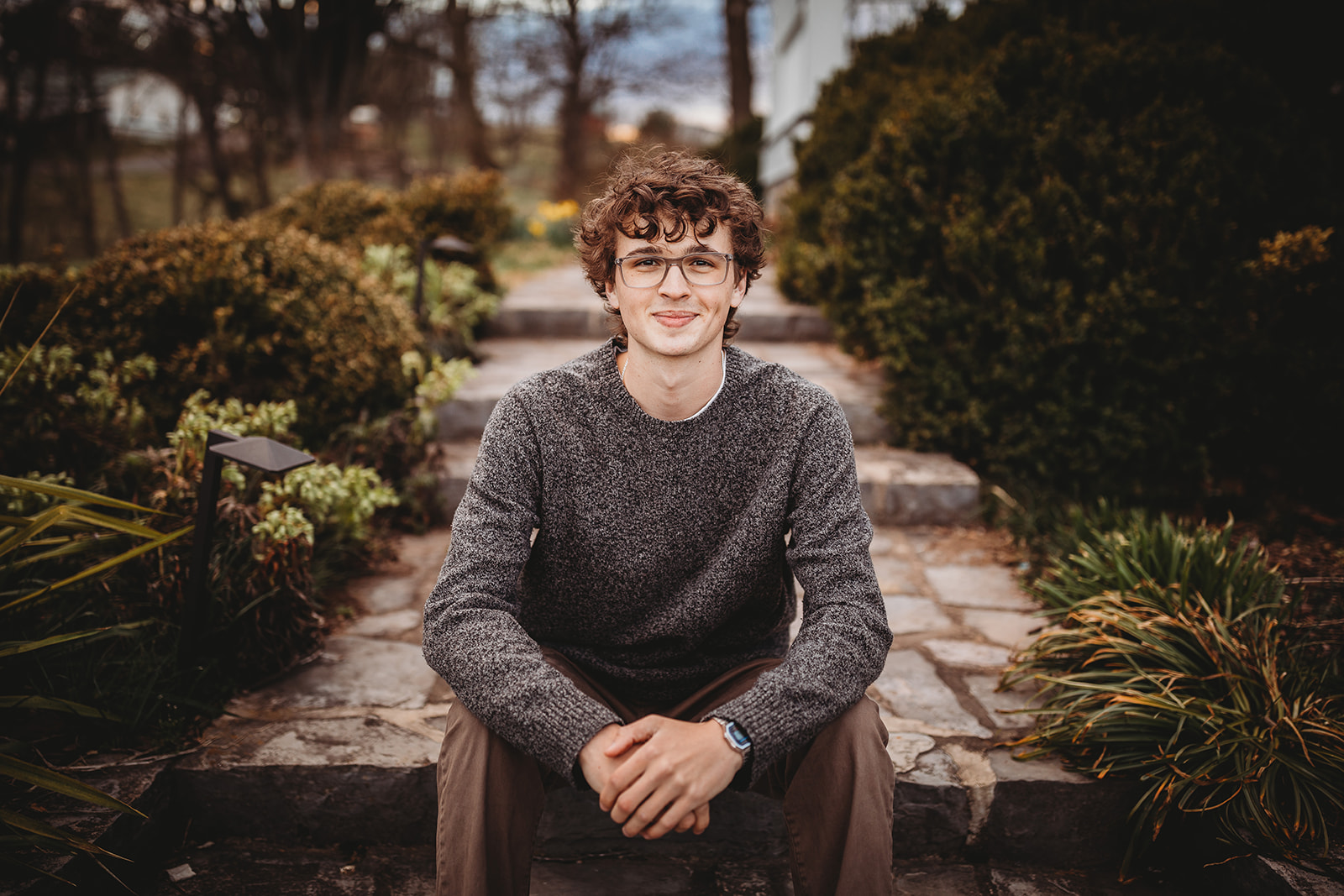 A high school senior in a grey sweater sits on stone garden steps leaning on his knees before finding Tuxedo Rental Harrisonburg, VA