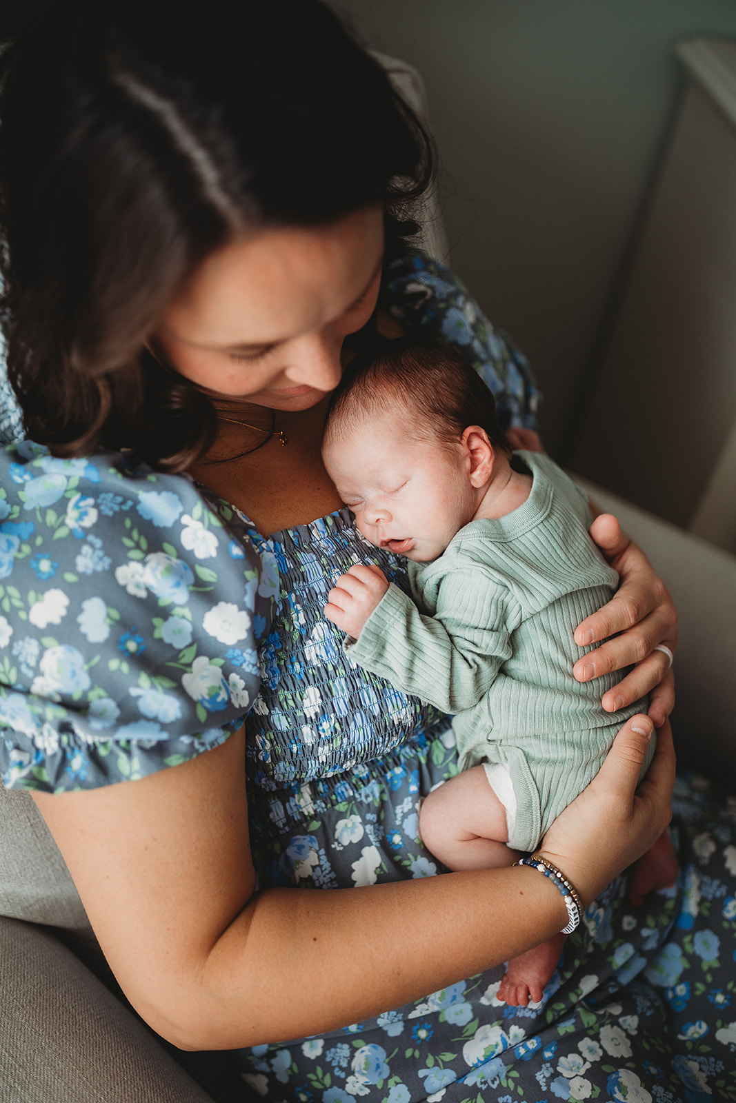 A newborn baby sleeps on mom's chest while sitting in a nursing chair under a window