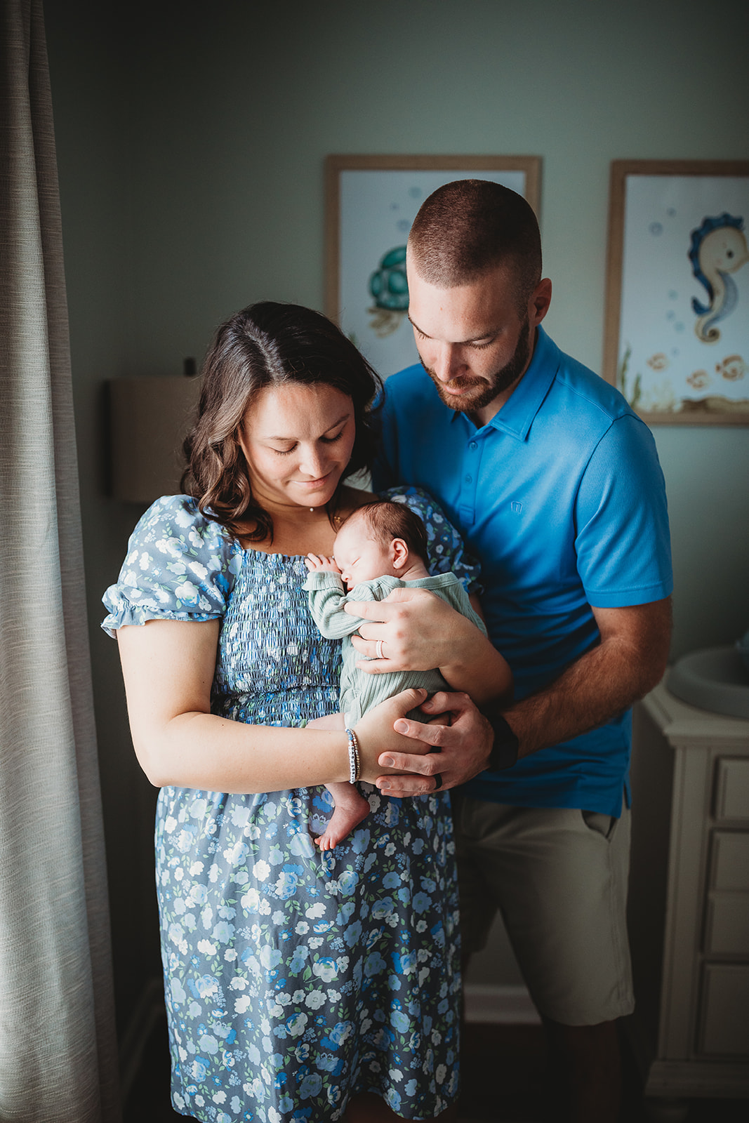 Happy new parents stand in a nursery window cradling their sleeping newborn baby