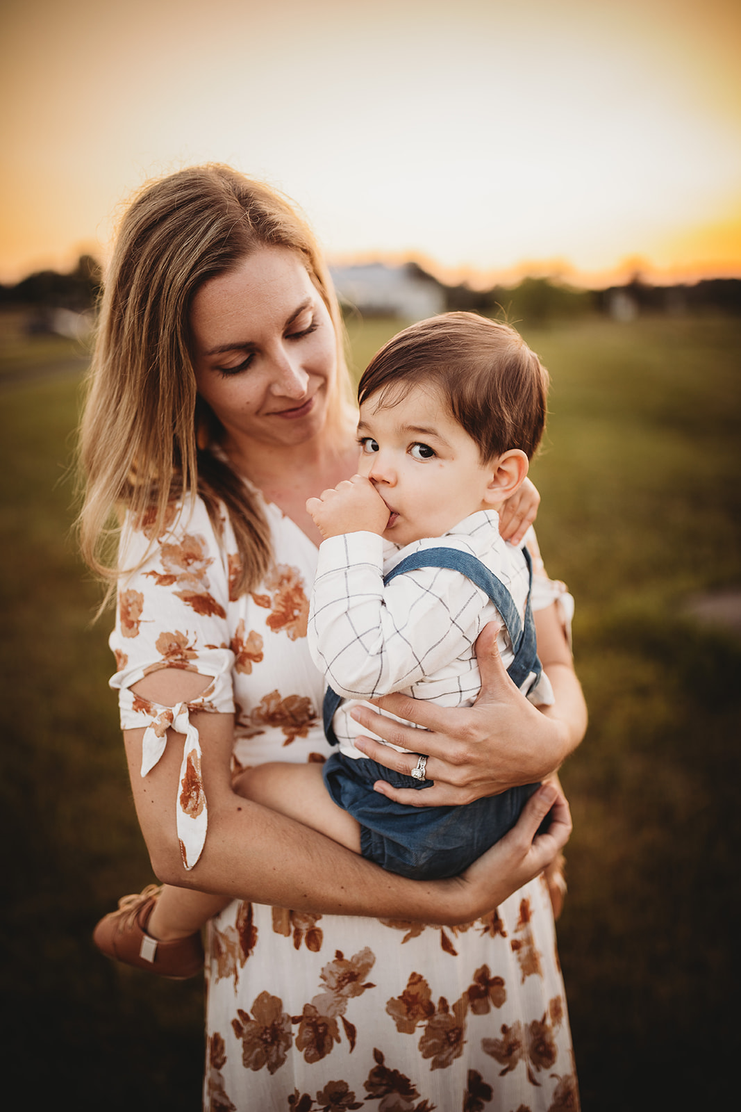 A toddler boy sucks his thumb while sitting in mom's arms in a field at sunset