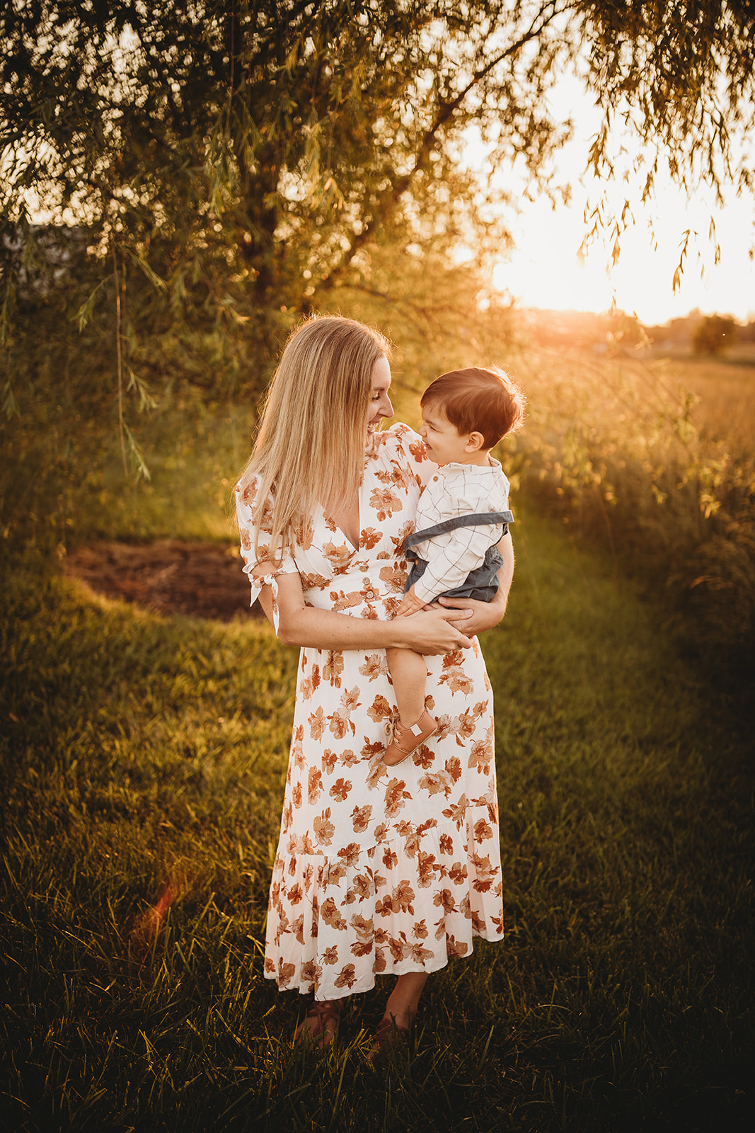 A happy mom smiles with her toddler on her hip in a field at sunset after visiting EMU Musikgarten