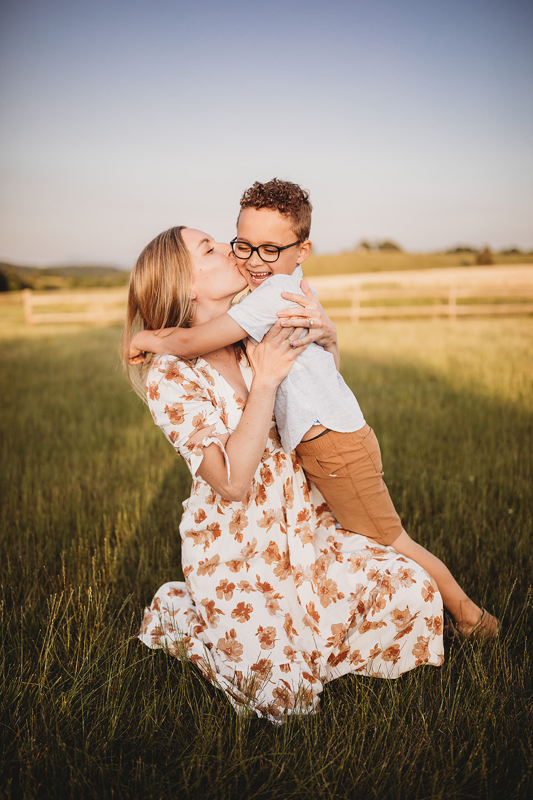 A toddler boy giggles while hugging and being kissed by mom in a field of grass after visiting EMU Musikgarten