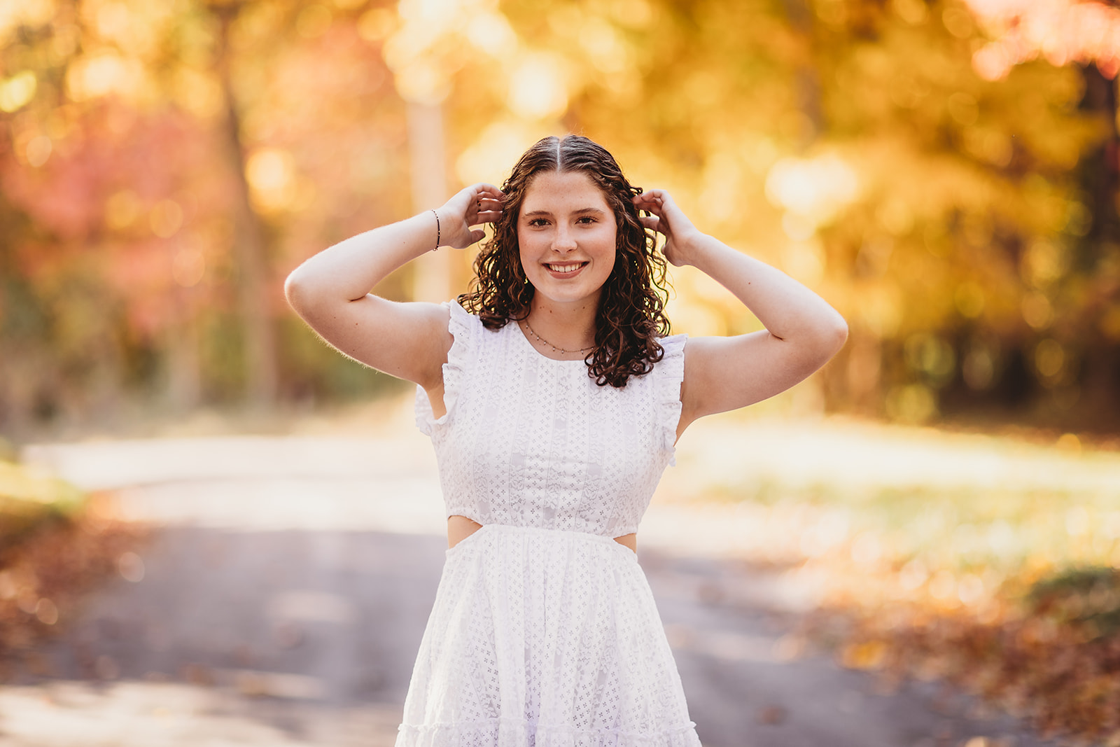 A high school senior puts her hands in her hair while walking on a street at sunset in fall