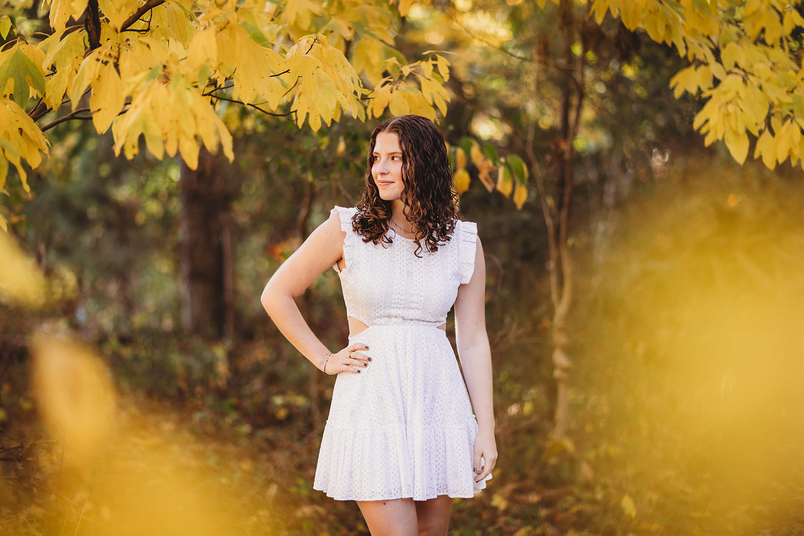 A woman in a white dress walks through fall colored trees at sunset with a hand on her hip