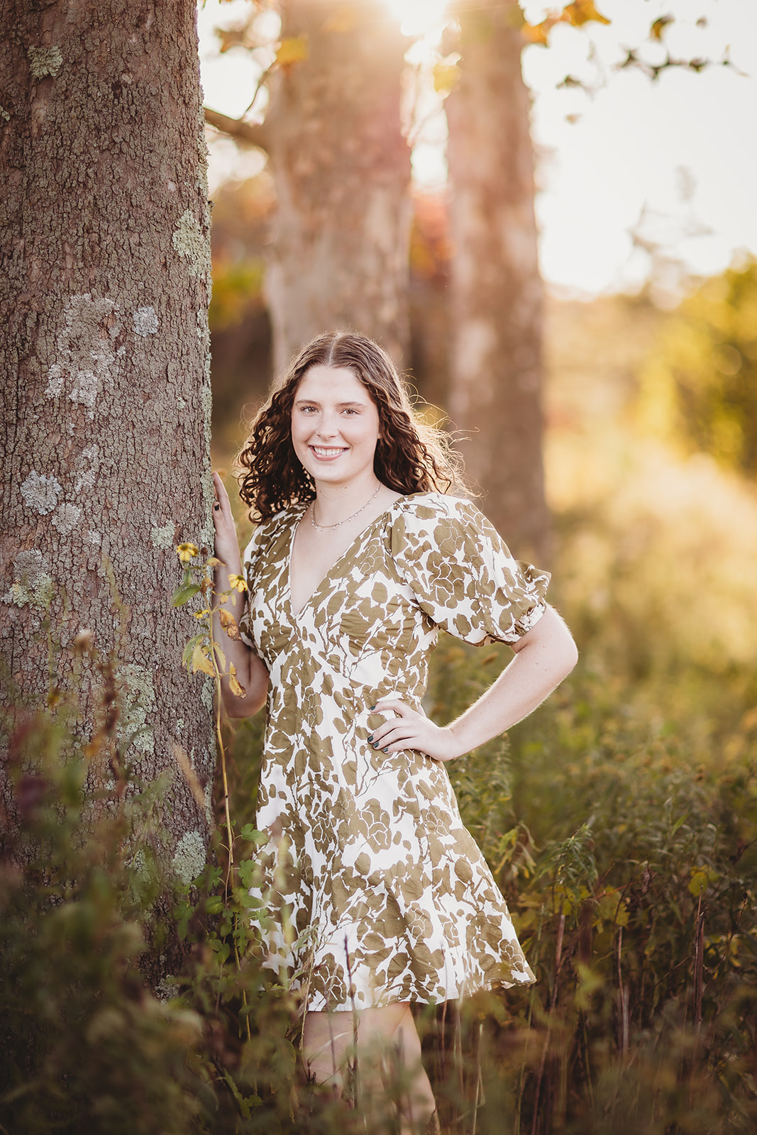 A woman in a brown floral print dress leans on a tree at sunset smiling after visiting Hair Salons in Harrisonburg, VA