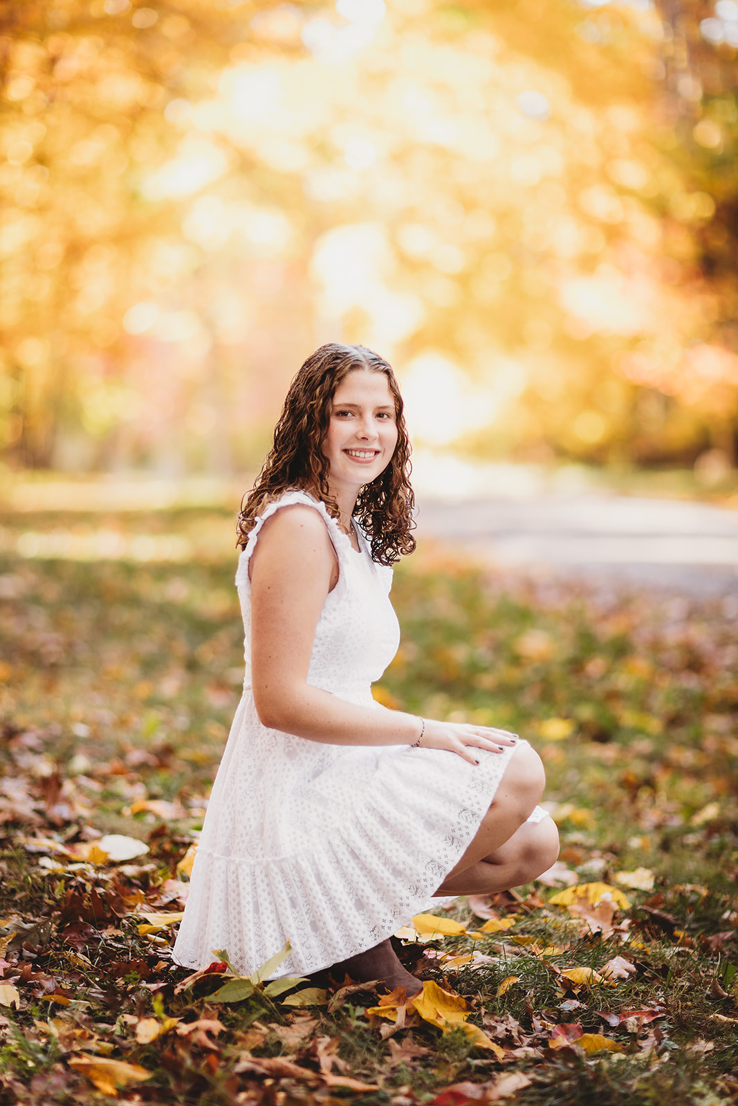 A high school senior in a white dress kneels in a park at sunset while smiling after visiting Hair Salons in Harrisonburg, VA