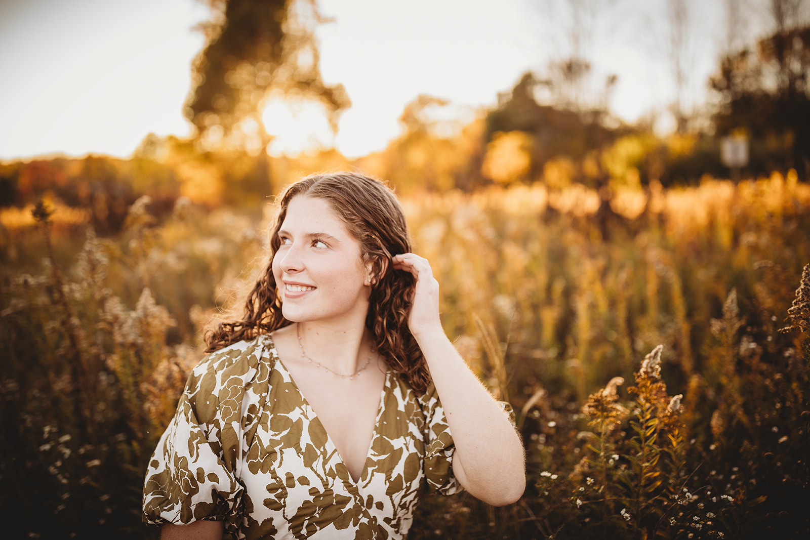 A woman in a floral print dress pulls her hair back while walking through tall golden grasses at sunset after visiting Hair Salons in Harrisonburg, VA