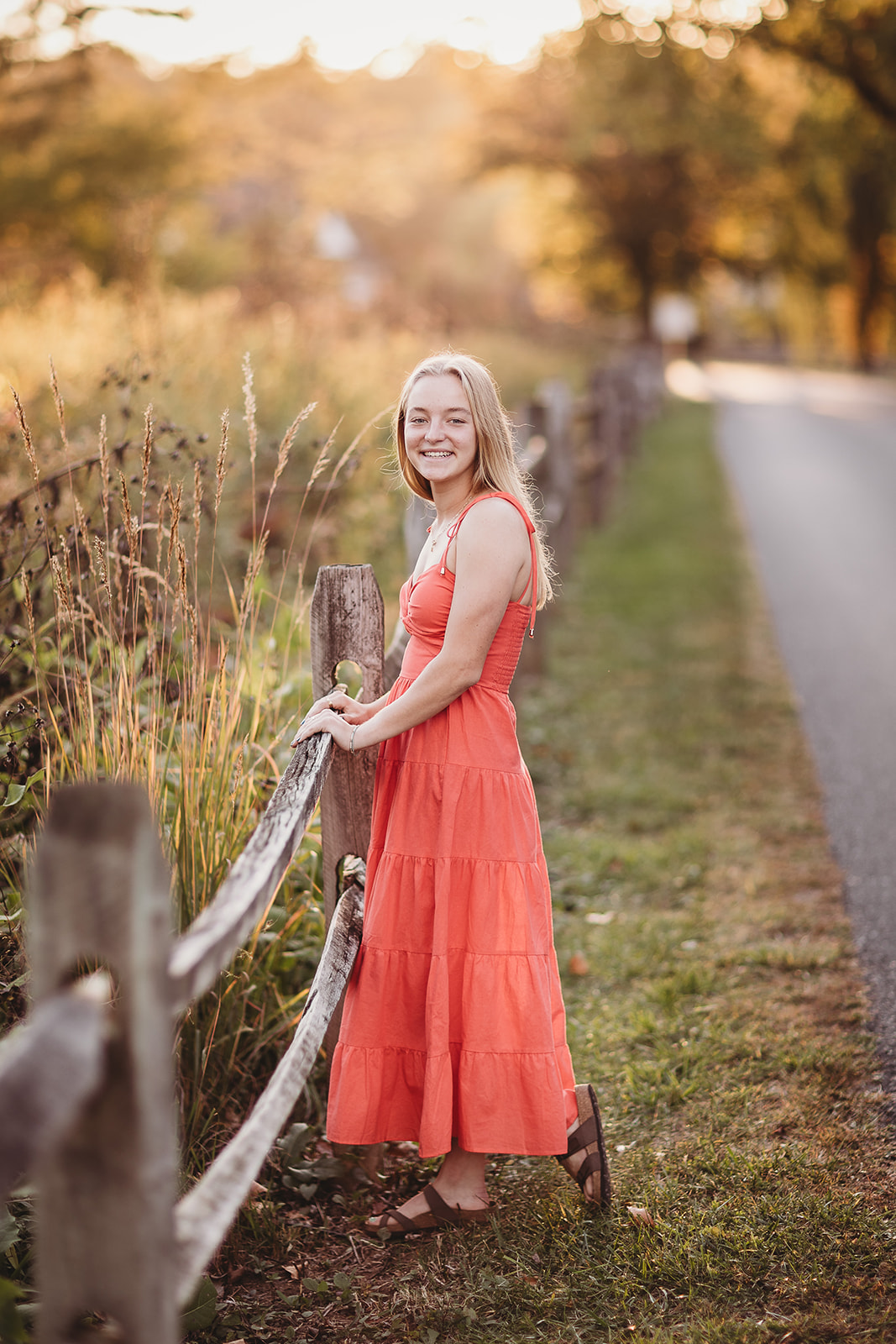 A smiling high school senior leans onto a wooden park fence by a sidewalk at sunset in a pink dress