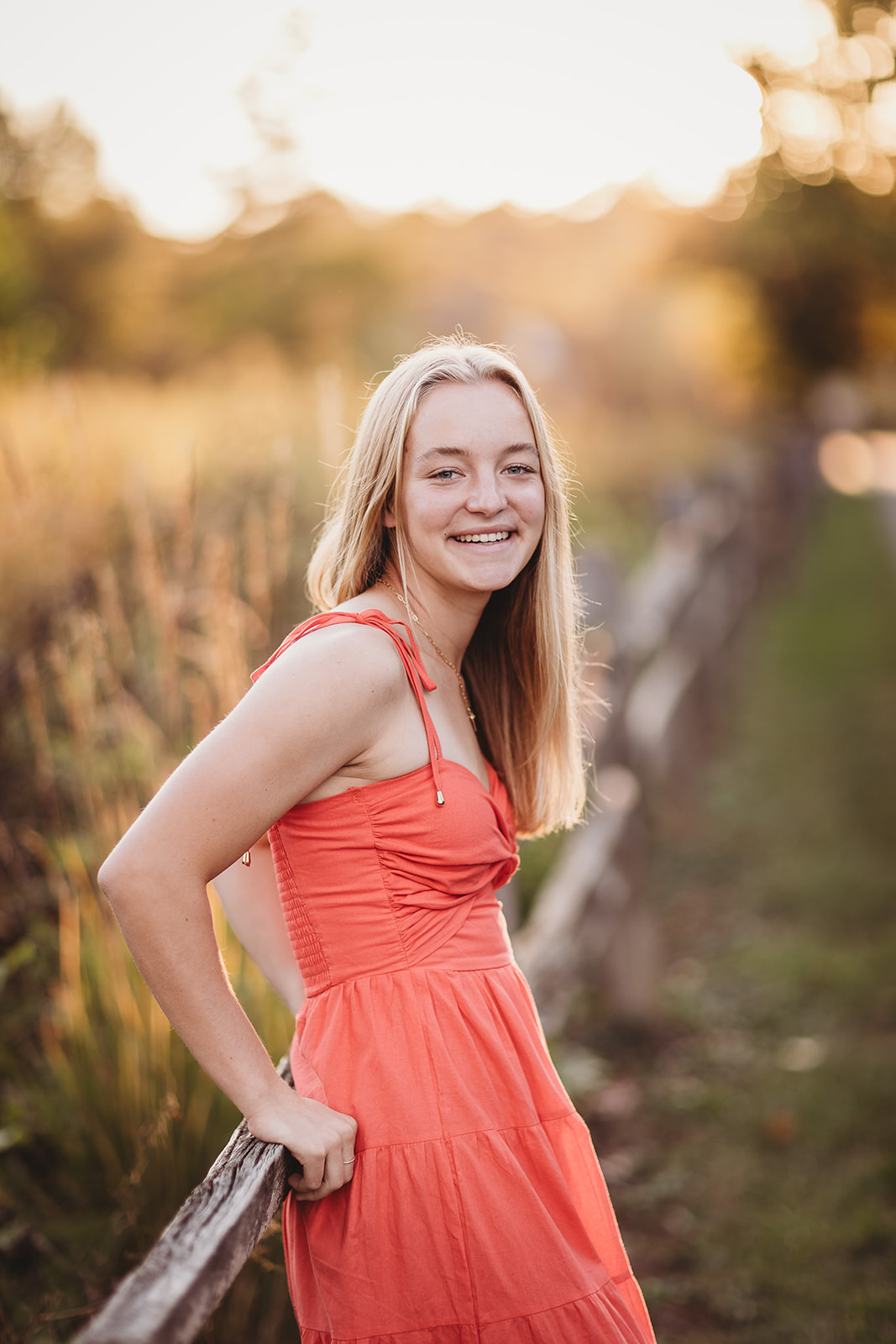 A smiling blonde high school senior leans on a wooden farm fence at sunset