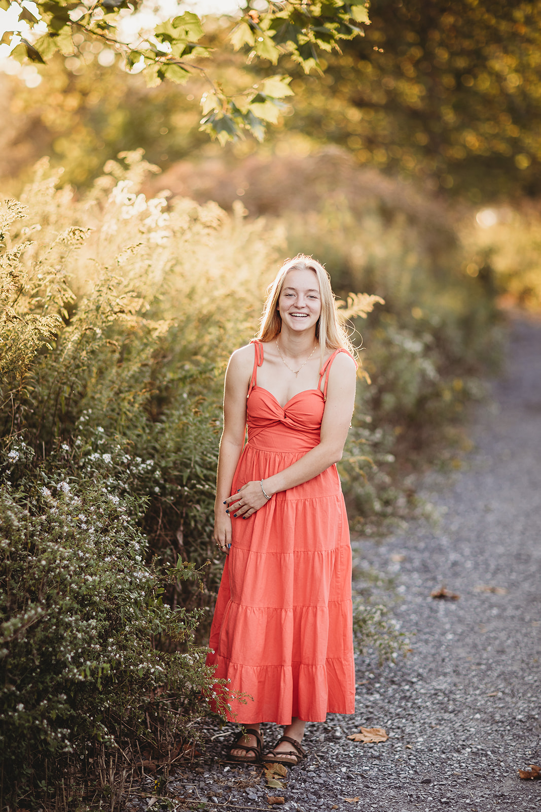 A high school senior in a pink dress laughs while walking a gravel trail at sunset after visiting Nail Salons in Harrisonburg, VA