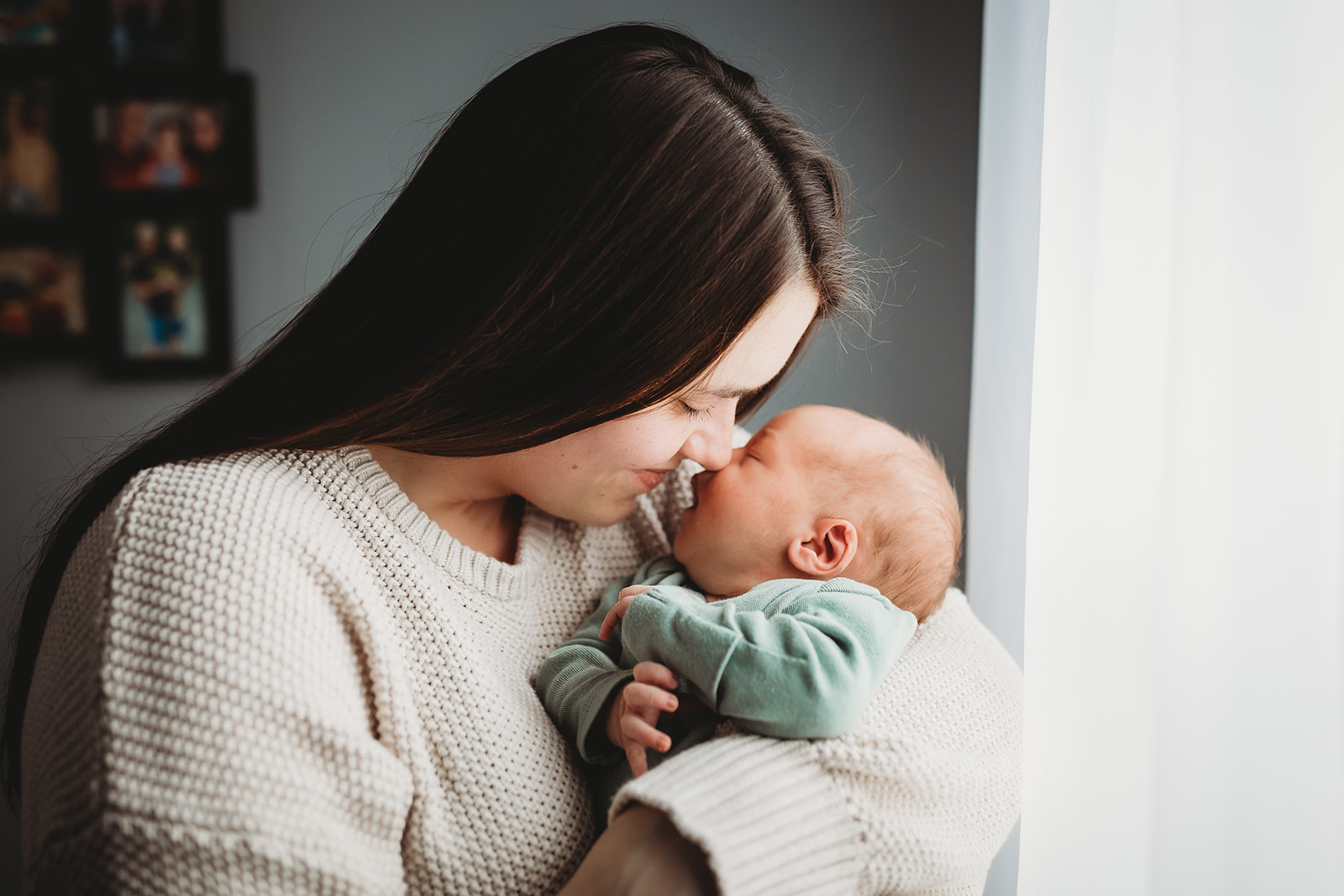 A mom snuggles her newborn baby while standing in a window in a sweater