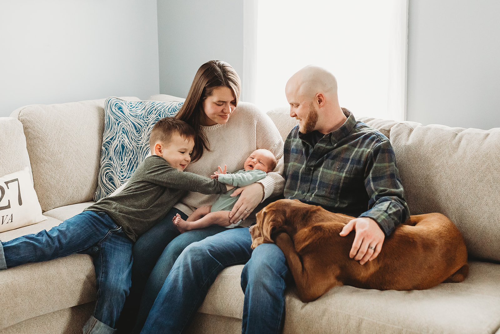 A happy mom holds her newborn baby on a couch after some Pelvic Floor Therapy In Harrisonburg, VA with dad, dog and toddler son