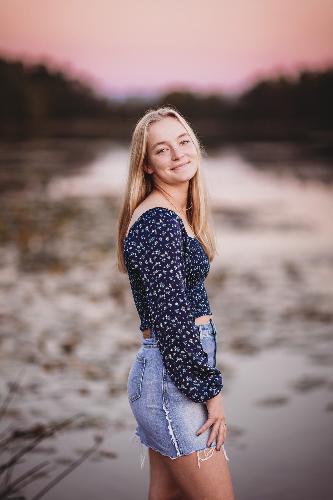 A happy girl in a floral blouse and jean skirt stands on a dock at sunset