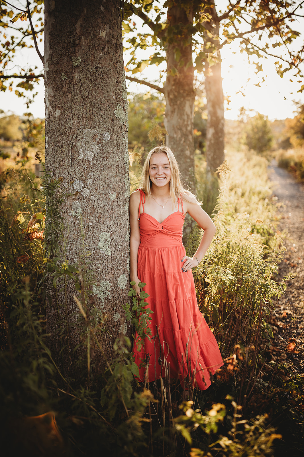 A high school senior leans on a tree in a trail at sunset in a pink dress after Tanning Harrisonburg, VA