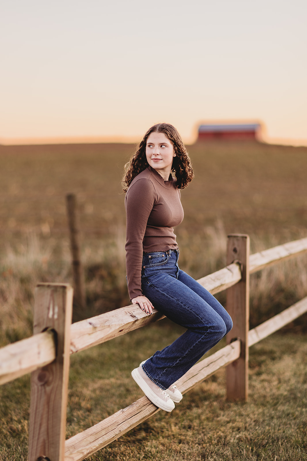 A high school senior in a brown shirt and jeans sits on a farm fence looking over her shoulder at sunset