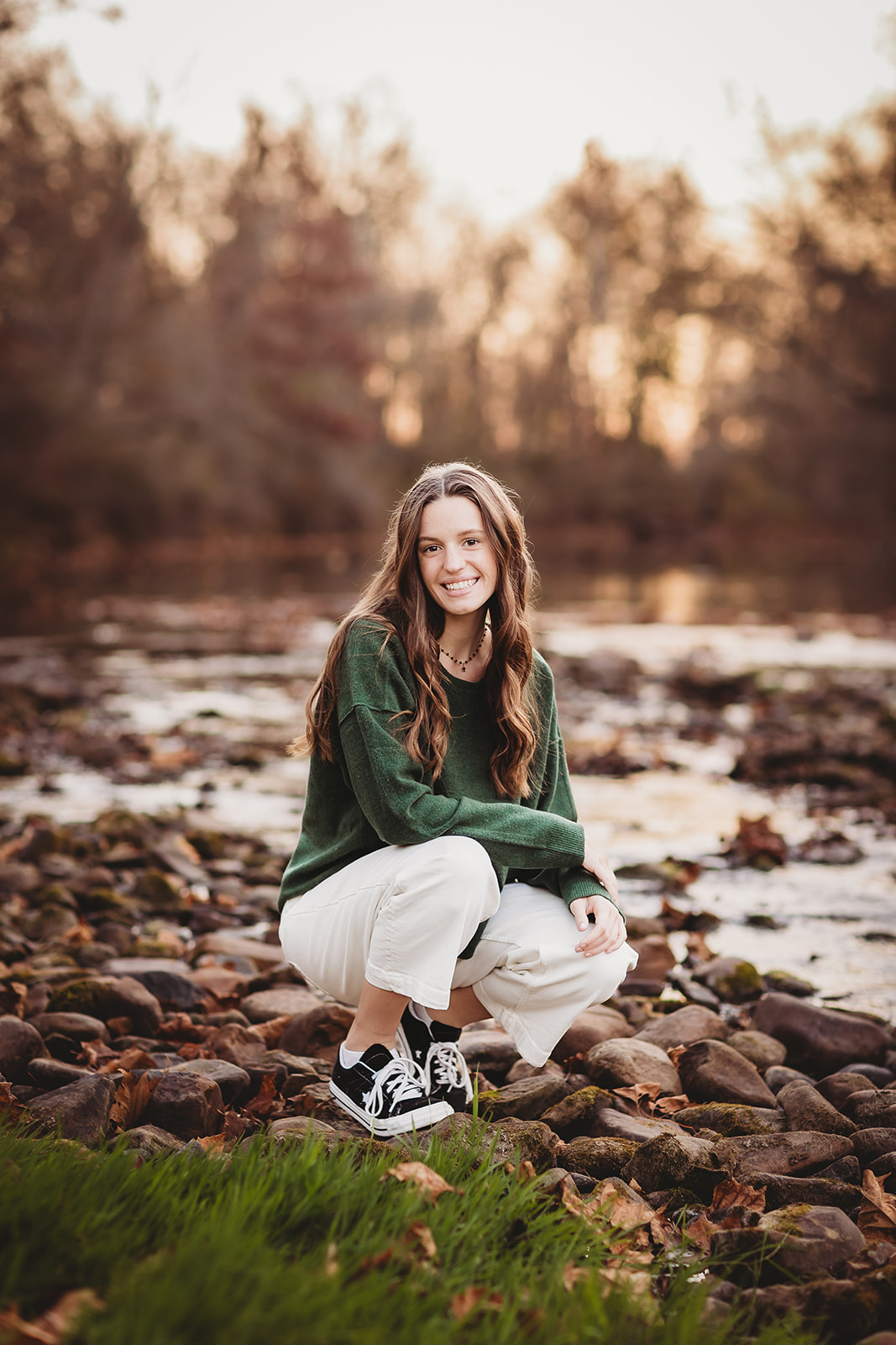 A woman in white pants and green shirt kneels by a creek on some rocks at sunset while smiling