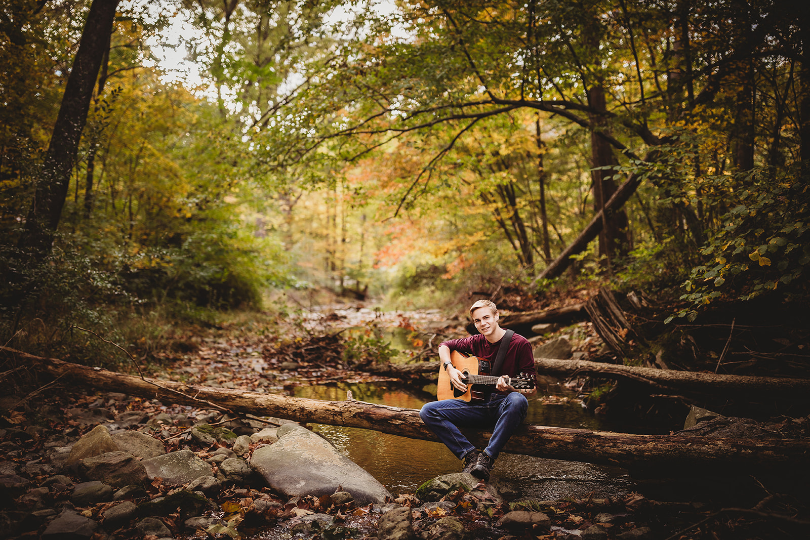 A high school senior boy in jeans sits on a log over a creek playing an acoustic guitar after exploring Volunteer Opportunities in Harrisonburg, VA