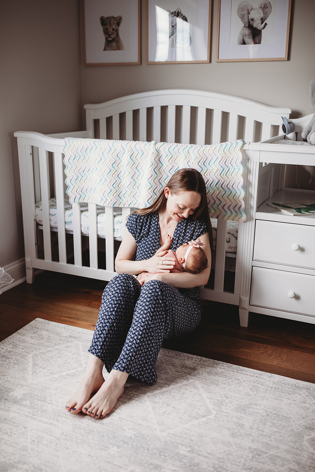 A happy new mom sits against a crib on the floor of a nursery cradling her newborn baby daughter