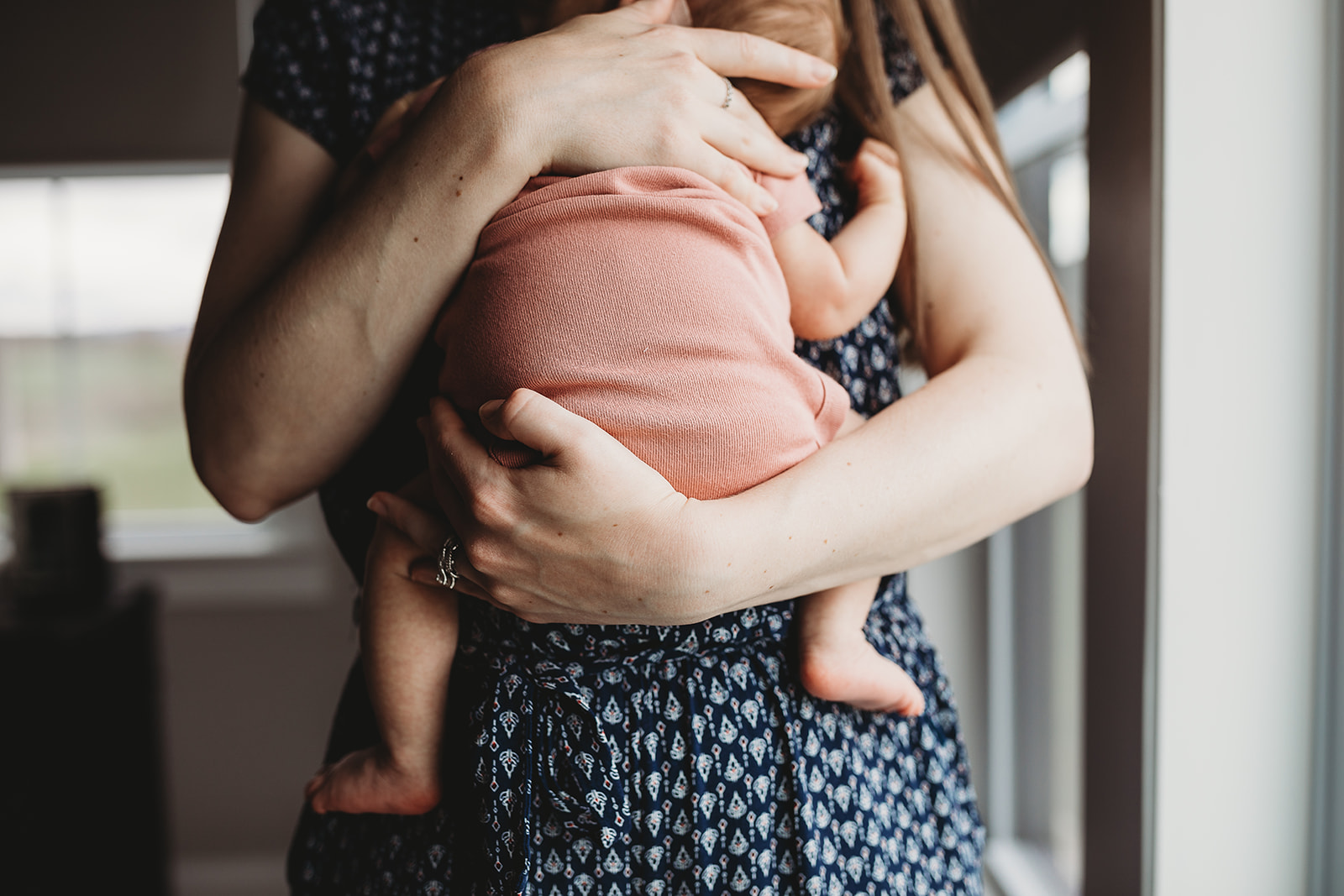 A newborn baby sleeps on mom's chest in her arms while mom stands in a window