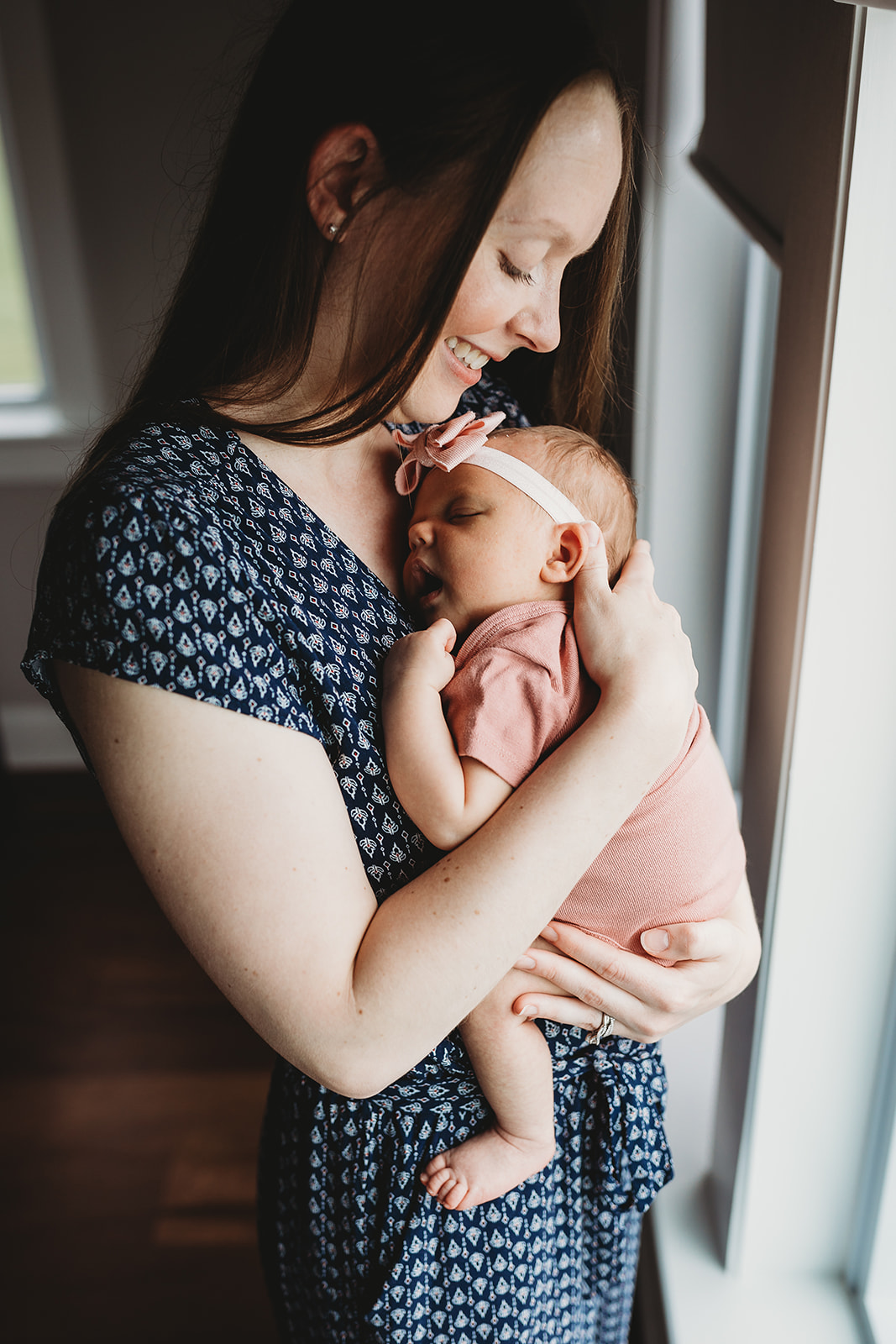 A happy mom snuggles her sleeping newborn daughter against her chest while standing in a window after some Harrisonburg Yoga