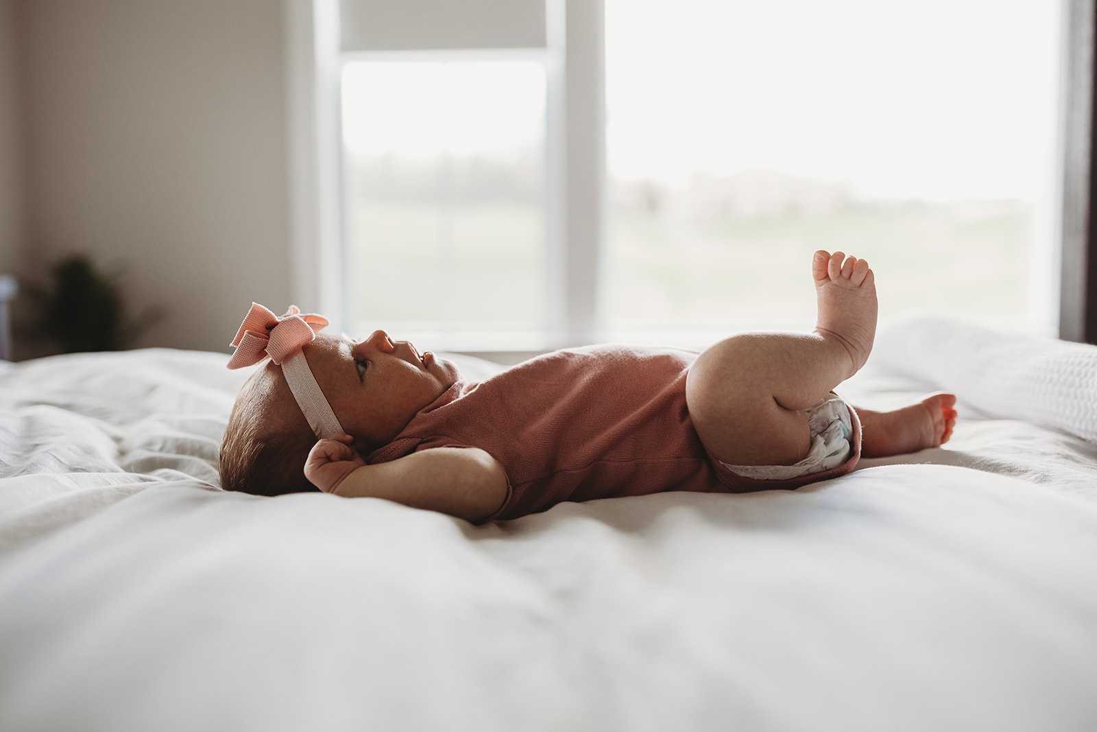 A newborn baby girl lays on a white bed under a window in a pink onesie and bow after mom did Harrisonburg Yoga