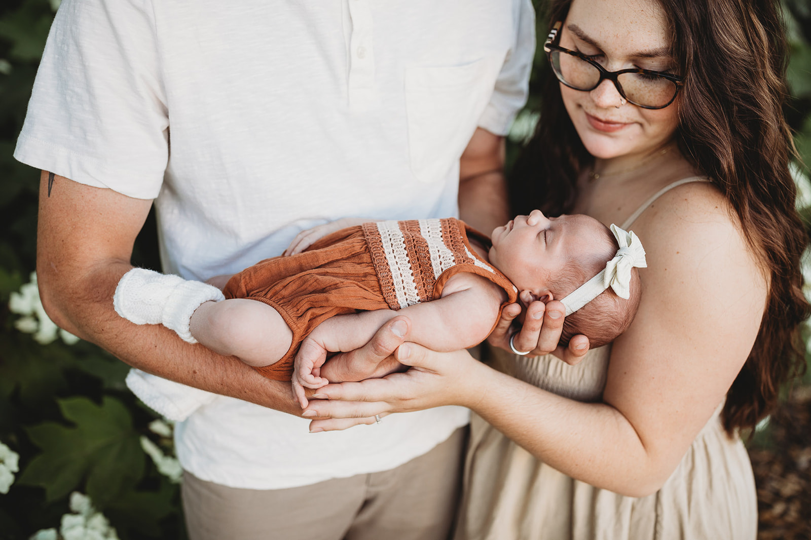 A newborn baby sleeps in dad's hands in an orange onesie and white bow